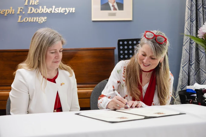 Two women sit at a table; one is signing a document while the other looks on. Both wear white jackets and red tops. Behind them is a piano and a sign that says "The Joseph F. Dobbelsteyn Lounge.
--
Deux femmes sont assises à une table ; l'une signe un document tandis que l'autre regarde. Toutes deux portent des vestes blanches et des hauts rouges. Derrière elles, un piano et un panneau indiquant "The Joseph F. Dobbelsteyn Lounge".