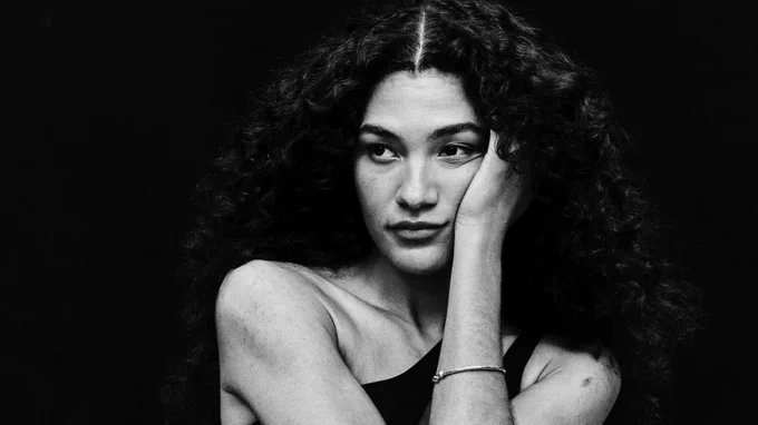 A black and white portrait of Olivia Dean with voluminous, curly dark hair. She is posing with one arm crossed over her chest and the other hand resting against her cheek. She wears a dark, one-shoulder dress with a sheer midriff panel and a pleated white skirt.