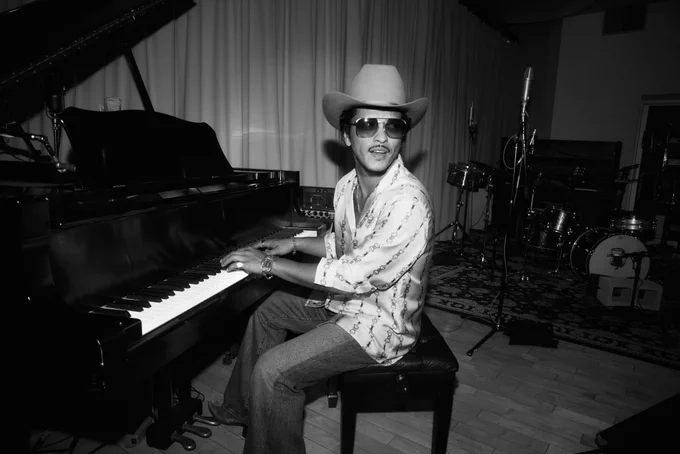 A black and white photograph of Bruno Mars sitting at a grand piano in a studio. He is wearing a patterned button-down shirt, jeans, sunglasses, and a wide-brimmed cowboy hat. He is looking over his shoulder away from the piano.