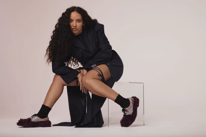Image of Jade with long, wavy dark hair. She is seated on a transparent acrylic cube against a plain, light-colored background. She is wearing a structured, navy blue blazer-style garment with long sleeves and sharp shoulders. 