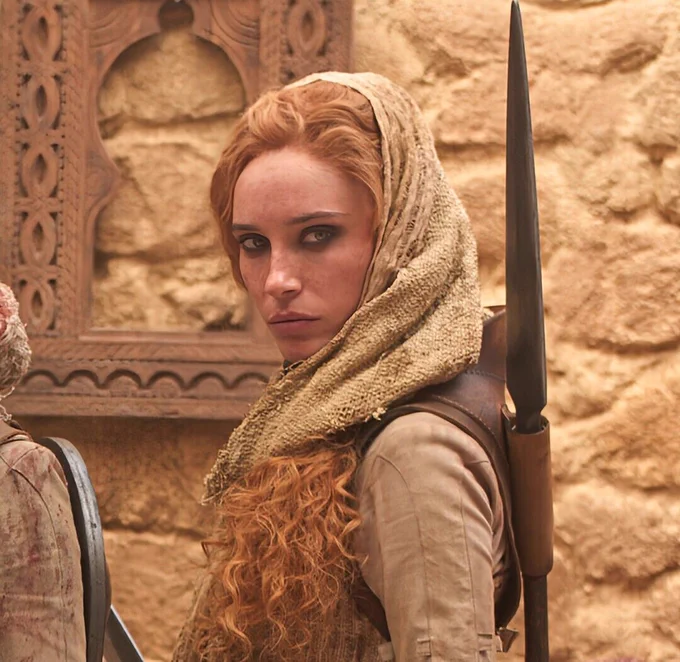 Maja Simonsen as Chiad in The Wheel of Time, wearing Aiel-style clothing with a light brown headscarf. She stands in profile against a stone wall, her long red hair visible beneath the scarf, with a spear secured on her back and a focused, watchful expression.