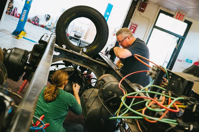Two mechanics working on a large truck in a workshop, one inspecting the wheel assembly and the other examining undercarriage components.