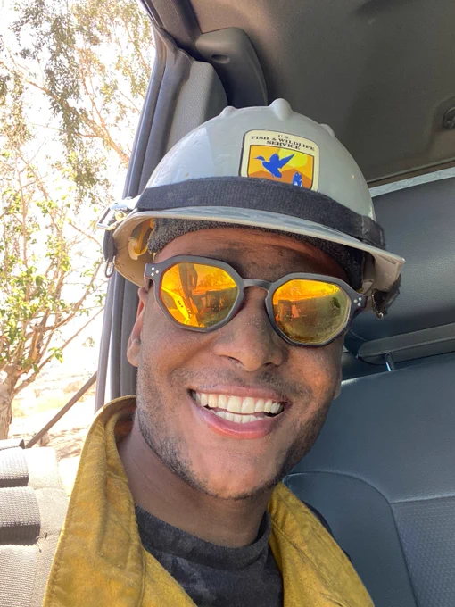 Firefighter smiling, wearing a hard hat and sun glasses inside a vehicle with trees visible outside.