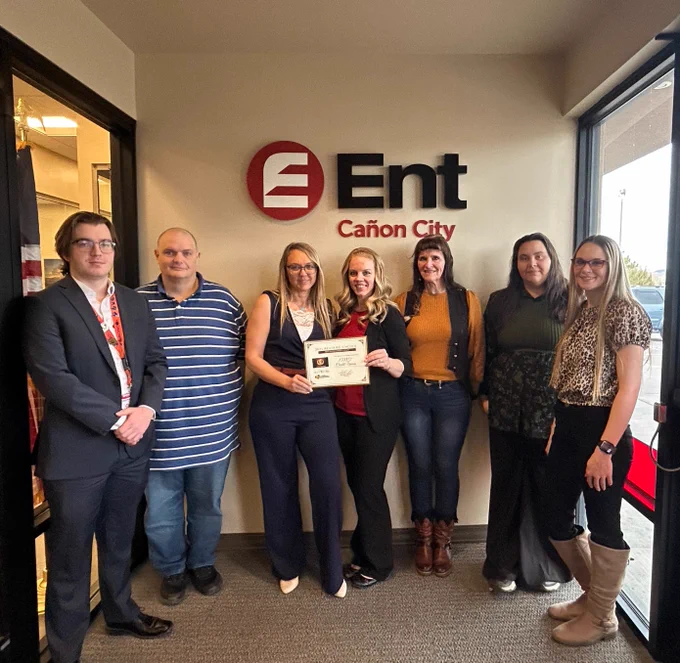 Group of seven people standing in a lobby with a sign reading "Ent Canon City" on the wall behind them. One individual is holding a framed certificate.