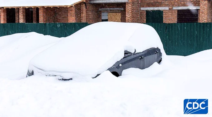 A car almost completely covered in snow, parked near a brick building.