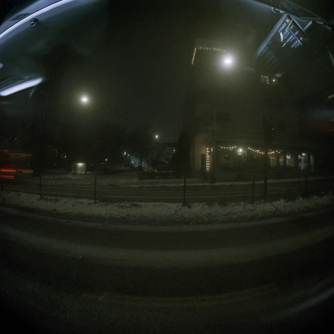 View from a bus window at night. Snow on the road, buildings, trees and lights further away.