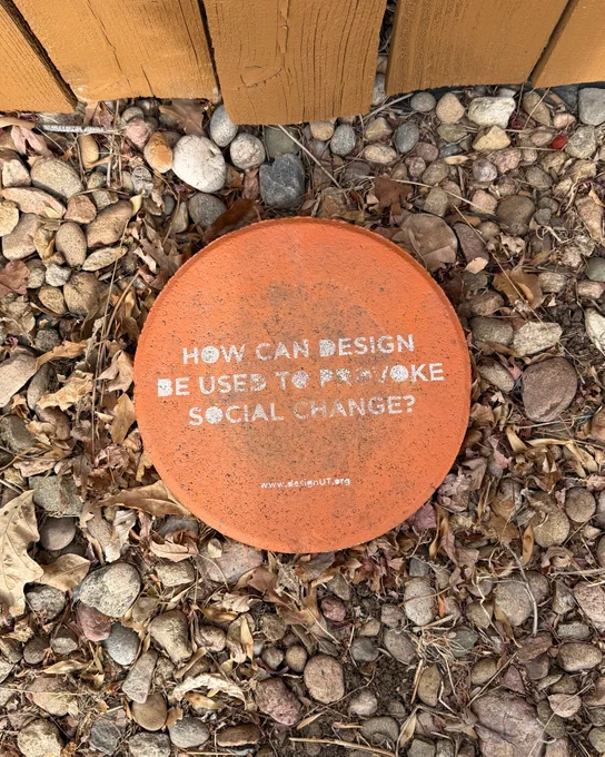 A painted orange round stone paver with white text on top that says, "How can design be used to provoke social change?"
