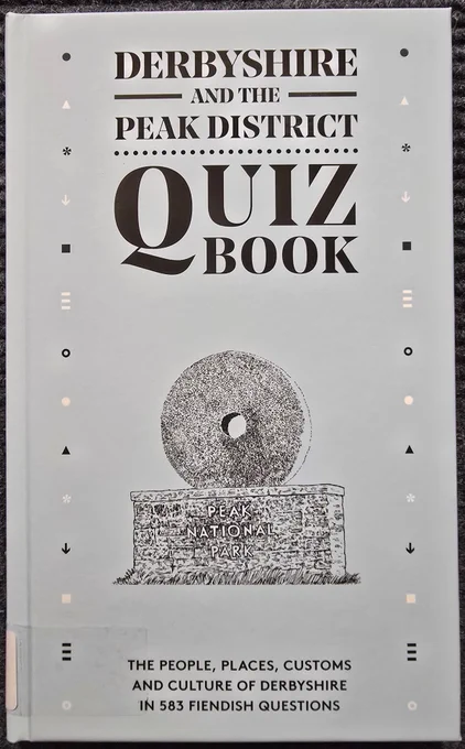 A hardback book in light blue with bold black title lettering in capitals reading' Derbyshire and the Peak District Quiz Book'. Below the title is a hand drawn black and white image of the Peak National Park symbol, a mill stone balanced on a stone plinth.
