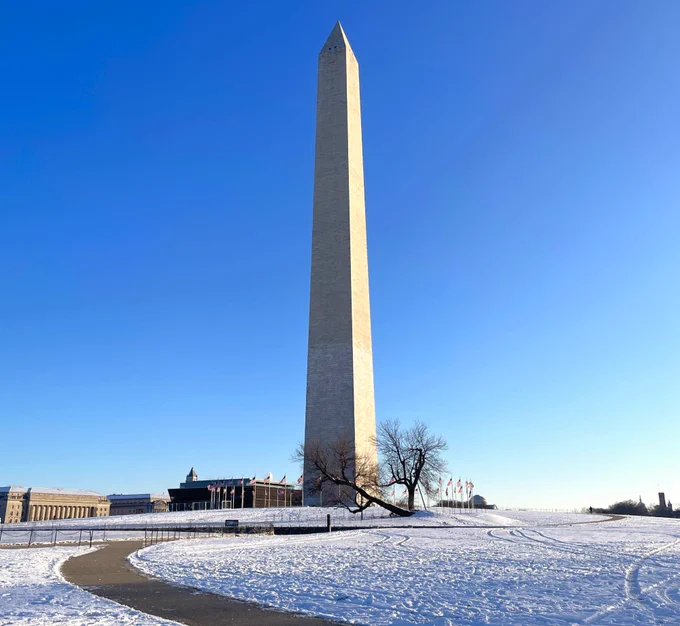 The tall white stone tower of the Washington Monument stands on a low snow-covered hill surrounded by a ring of American flags under a bright blue sky.