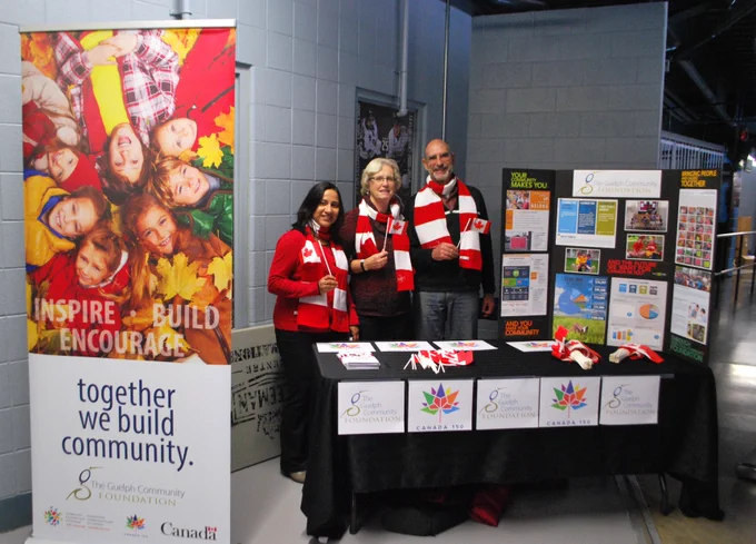 Three people standing together behind a booth at the hockey arena with red and white scarves and with the GCF signs
