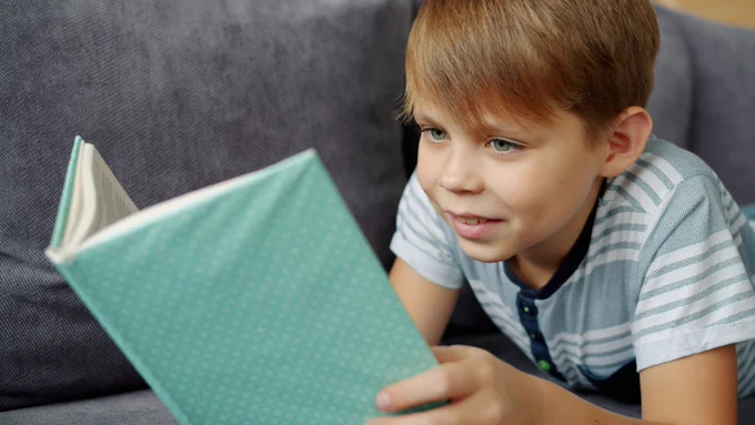 A young boy with light brown hair and a striped shirt is lying on a gray couch, smiling while attentively reading a teal book.