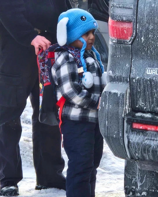 A photo of Liam outside wearing a hat and a backpack. Someone is holding him by his backpack.