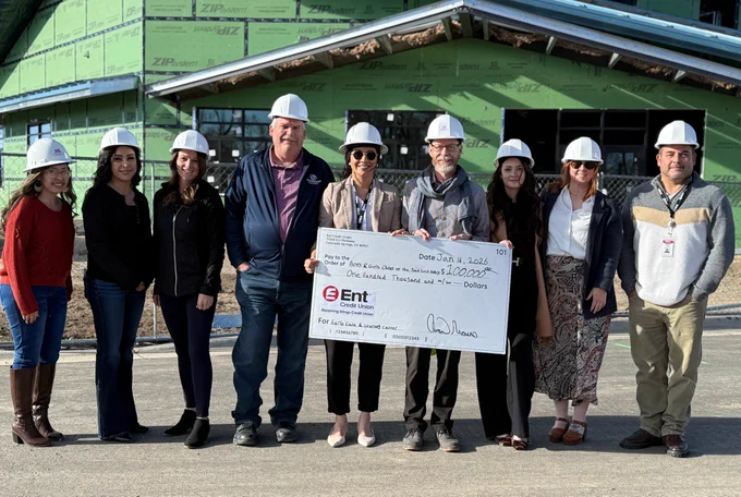 Group of people in hard hats holding a large ceremonial check from Ent Credit Union in front of a construction site.