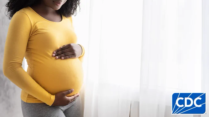 Image shows a close-up of a pregnant individual in a yellow top, holding their belly gently, standing next to a window. 