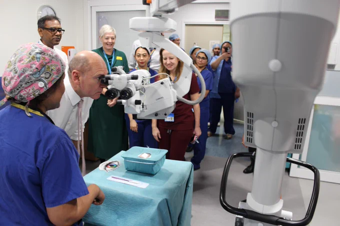 Hospital staff and Sir Jim Mackey observe a dummy surgical demonstration in a hospital room with advanced equipment.