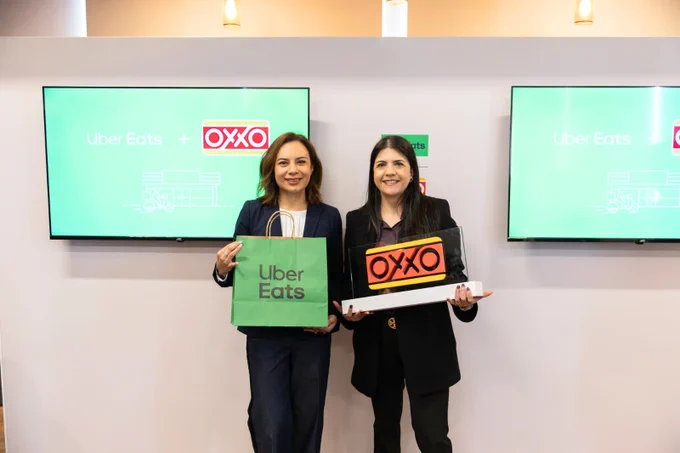 Dos representantes de OXXO y Uber Eats posan sonrientes para celebrar su alianza estratégica en México. La representante a la izquierda sostiene una bolsa de Uber Eats, mientras que la de la derecha sujeta una placa con el logotipo de OXXO, simbolizando la integración de ambas marcas para ofrecer conveniencia a domicilio.