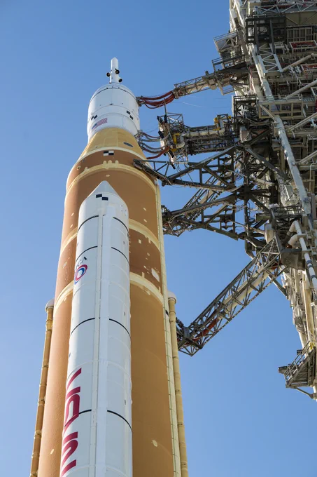 NASA\u2019s Artemis II Space Launch System (SLS) rocket and Orion spacecraft, secured to the mobile launcher, are seen as they make the 4.2 mile journey toward Launch Pad 39B, Saturday, Jan. 17, 2026, at NASA\u2019s Kennedy Space Center in Florida. The perspective of the image is from below the rocket looking up. The core stage of the rocket is orange and topped with the white Orion spacecraft and launch abort system. There is a white solid rocket booster on the side of the rocket facing the viewer. The sky is blue in the background. Credit: NASA/Aubrey Gemignani