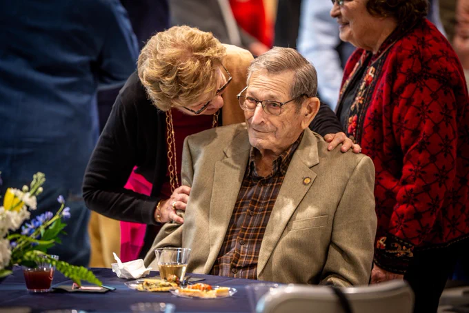One individual sitting at a table while another leans down to talk to them. 