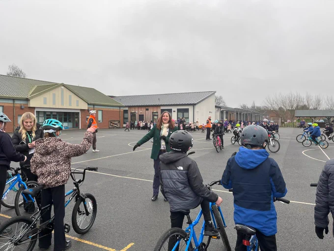Children wearing helmets riding bicycles in a school playground with an instructor guiding them on a cloudy day.