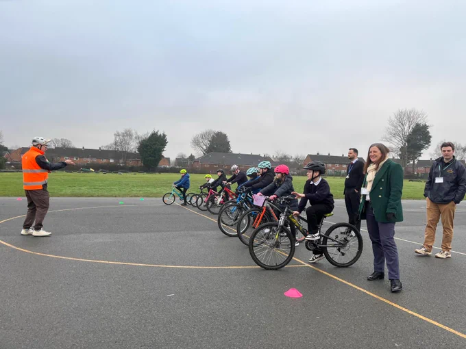 Group of children wearing helmets lined up on bicycles in a playground, receiving instructions from an adult in an orange vest.