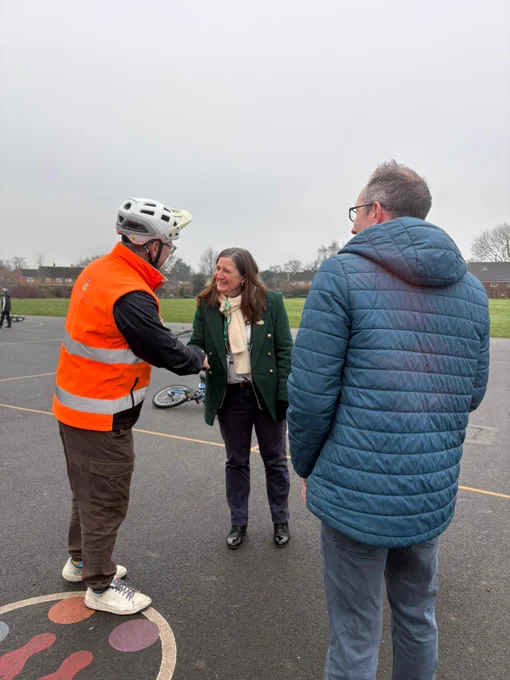 Person in orange safety vest and helmet shaking hands with a woman on an outdoor basketball court on a cloudy day.