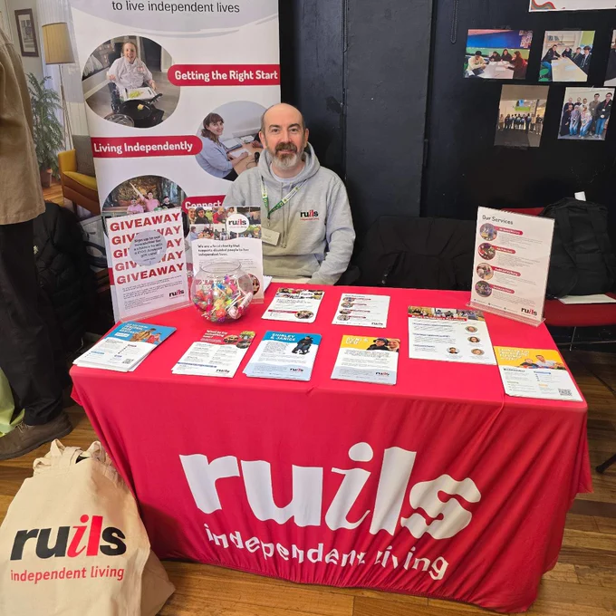 A person sitting behind a Ruils Independent Living information table with brochures and giveaway items at an indoor event.