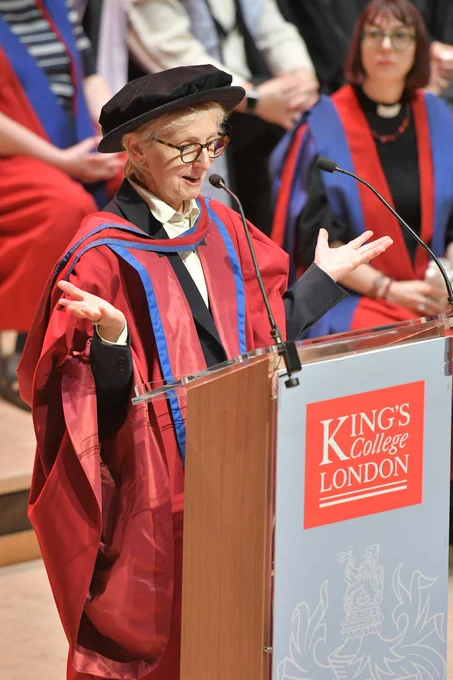 A person in red graduation robes stands at a lectern delivering a speech, hands aloft.