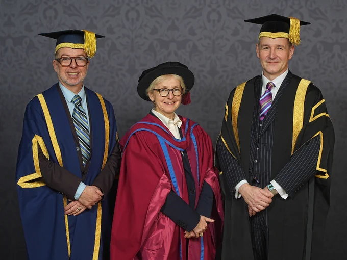Three people pose for a graduation photo; a shorter person in the middle wears red robes and a black cap, flanked by two people in black and yellow-trimmed robes wearing mortarboards.
