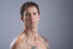 Shirtless man with short hair and visible tattoos on his shoulder and chest against a plain gray background.