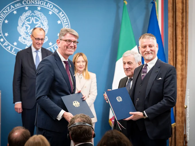 Wolfram Weimer und sein Amtskollege Alessandro Giuli präsentieren zwei Mappen mit Abkommen, die sie für den Kulturbereich vereinbart haben. Im Hintergrund stehen Bundeskanzler Friedrich Merz und Regierungschefin Giorgia Meloni.