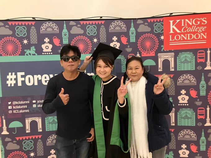 A graduate in a green and black gown posing for a picture with friends and family in front of the forever King's sign.