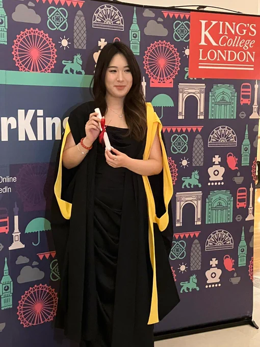 A graduate in a yellow and black gown, holding a graduation scroll, posing for a picture with friends and family in front of the forever King's sign.