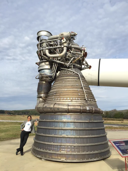 A woman leans against a large rocket engine on a sunny day. 