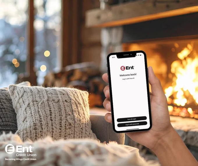 Person holding a smartphone displaying the Ent Credit Union login page, with a cozy fireplace in the background.