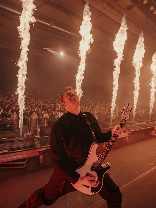 Ein Mann auf der Bühne spielt eine elektrische Gitarre, während im Hintergrund Feuereffekte brennen.