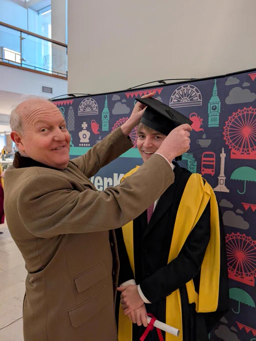 A parent playfully putting a graduation hat on a graduate who is dressed in a yellow and black academic robe.