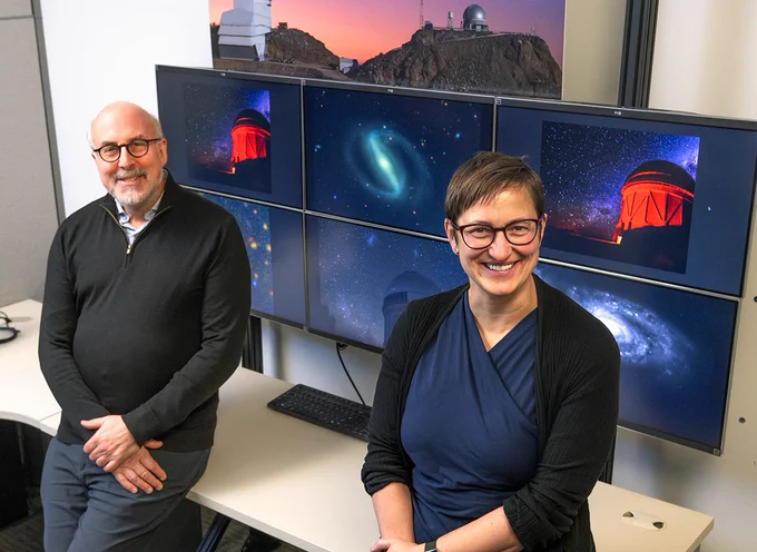 Josh Frieman and Risa Wechsler in the Rubin Control Room at SLAC. Six computer monitors behind them show images of Rubin Observatory and various cosmic phenomena. (Jacqueline Orrell/SLAC National Accelerator Laboratory)
