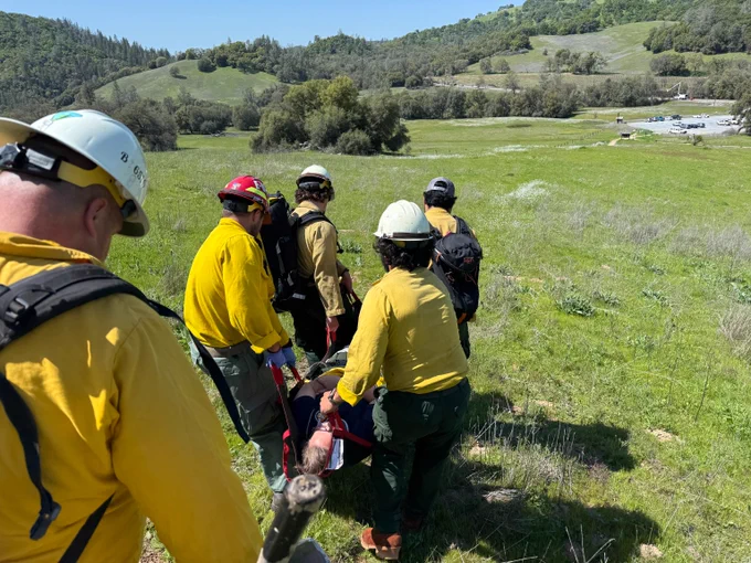 Firefighters in nomex carry an injured person on a stretcher across a grassy field with hills in the background during a training. 