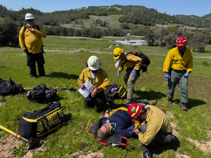 Firefighters in nomex attending to a person lying on grass in a rural field during a rescue drill.