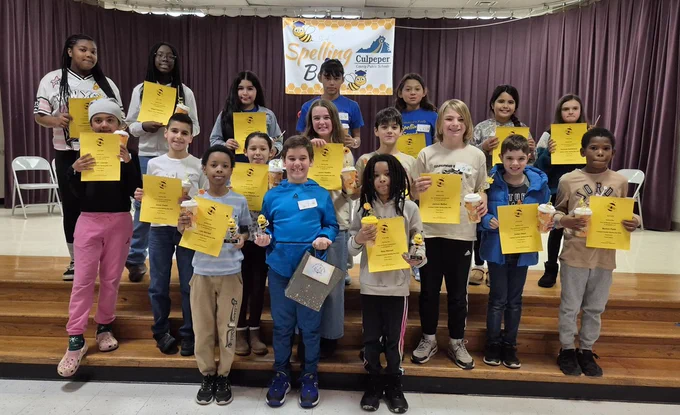 group of elementary students on a school stage holding certificates.  there is a sign above them that says CCPS Spelling Bee in front of a purple curtain