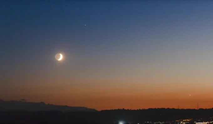 Esta imagen muestra una luna creciente brillando en el cielo durante el atardecer. Saturno se encuentra m\u00E1s arriba y a la derecha de la Luna. El cielo muestra un degradado de colores, del naranja cerca del horizonte al azul oscuro en la parte superior. En la parte inferior se distinguen siluetas de colinas y luces dispersas de una zona urbana.\n 