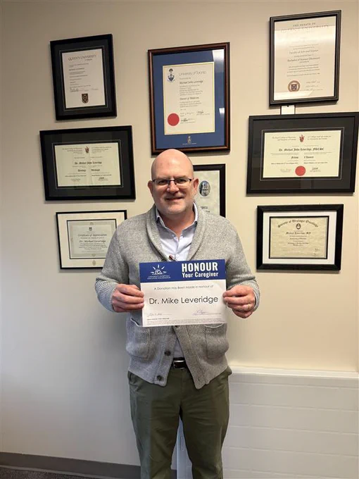 Person holding an 'Honour Your Caregiver' certificate, standing in front of a wall adorned with various framed certificates, including one labeled 'Dr. Mike Leveridge'.