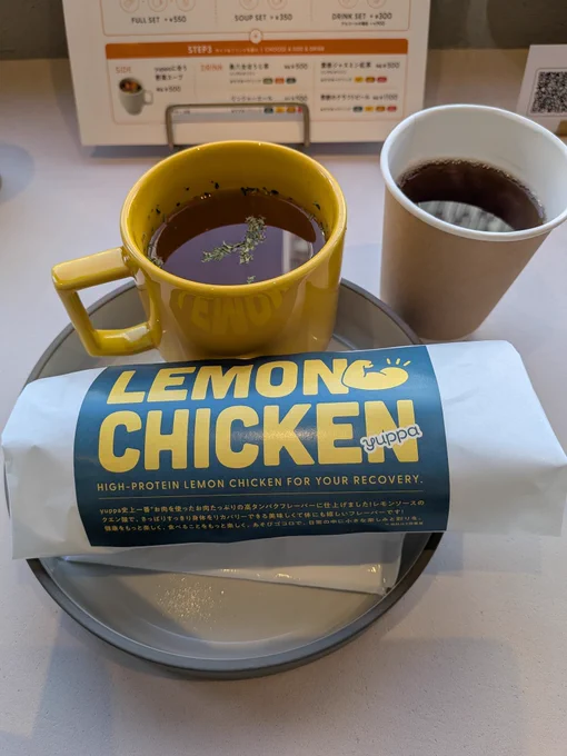 丸くて深いお盆に載せられた包みとスープのカップ。その右上に紙カップのお茶。包みには「LEMON CHICKEN yuppa」と描いてある。