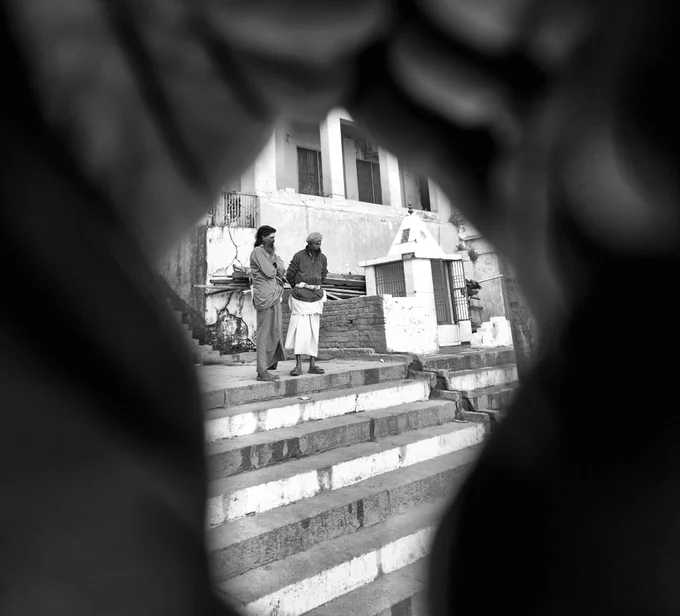 A black and white street photograph featuring a high-contrast 'keyhole' composition. The foreground is dominated by a dark, out-of-focus frame (likely created by hands ), directing focus to the center. There, two men in traditional attire stand on stone steps next to a small temple structure, set against a backdrop of aging buildings. The image creates a voyeuristic feel, isolating the subjects in their environment.