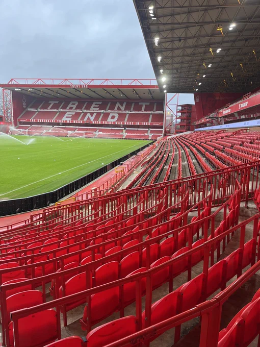 Photo of an empty City Ground, home of Nottingham Forest FC. 
The shot is taken at the very end of the Brian Clough stand lower tier, where it joins the Bridgford stand. The bottom third of image depicts curved rails and red seats which sweep round tho the right, joining the lower tier previously mentioned. Hovering above the lower tier a light grey roofing structure can be seen with a web of ironwork and some lighting in its underbelly. At the far top right of the photo you can just make out a stack of red shipping containers which are used as corporate suites, these are at the very opposite end of the Clough stand. 
Towards the top left of the image a bright green pitch is being watered by a series of automatic sprinklers in front of the double tier Trent End stand. The words “Trent End” are clearly visible in white seating in the top tier of this stand against the backdrop of the predominantly red seats. 
The photo was taken in daylight and shows a light blue/grey  sky above.