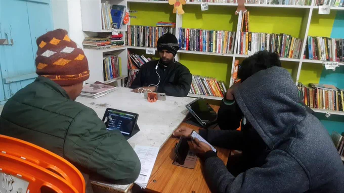 Four Bansa Community Library members sit around a table inside the library, using tablets and a mobile phone for study and preparation, with bookshelves filled with books in the background, highlighting access to digital learning alongside physical books.