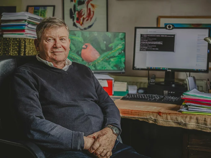 A man wearing a grey jumper with a shirt underneath sits in a chair at a desk. On the desk is a computer, a keyboard and several work files.