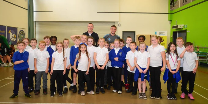 Group of children with two adults posing in a gymnasium, wearing sports and school uniforms.