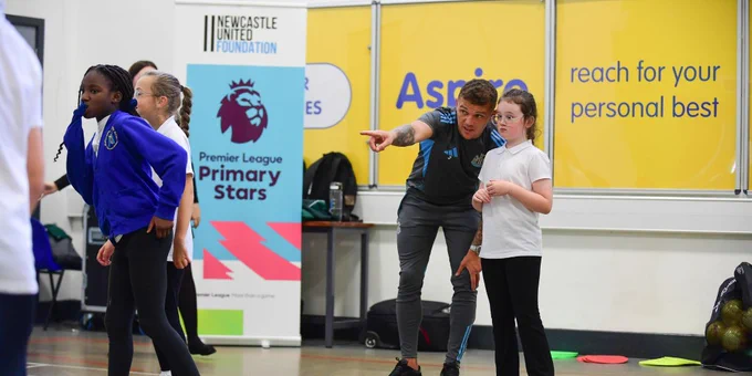 Children participating in a sports activity in a gym under banners of the Newcastle United Foundation and the Premier League Primary Stars programme.