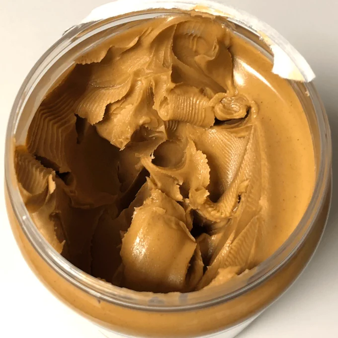 An open jar of creamy peanut butter viewed from above. The peanut butter is light brown with smooth, thick swirls and ridged scoop marks across the surface. The foil seal is partially peeled back and folded over the rim of the clear plastic jar. The jar sits on a light-colored surface with even lighting and soft shadows.

